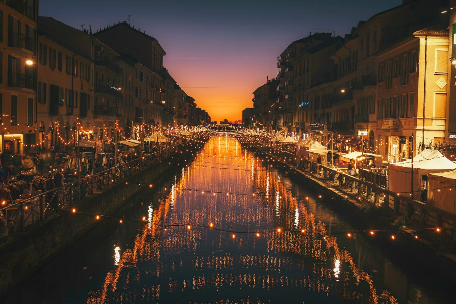 Navigli canal in Milan at blue hour with string lights, market stalls and shuttered apartment facades reflected in the water
