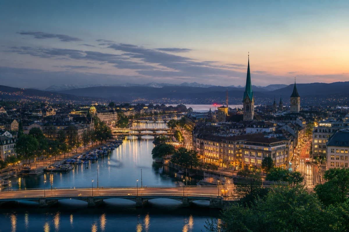 Zurich at dusk along the Limmat with banking-district lights and alpine foothills beyond